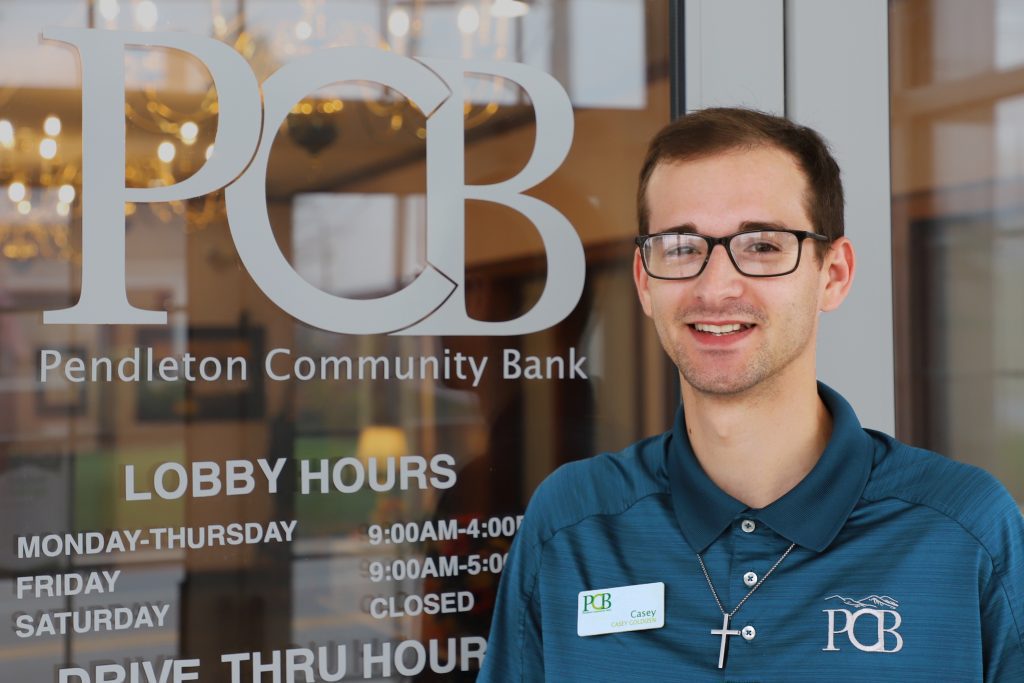 A man wearing glasses stands in front of a glass door that has lettering on the door:" PCB - Pendleton Community Bank."