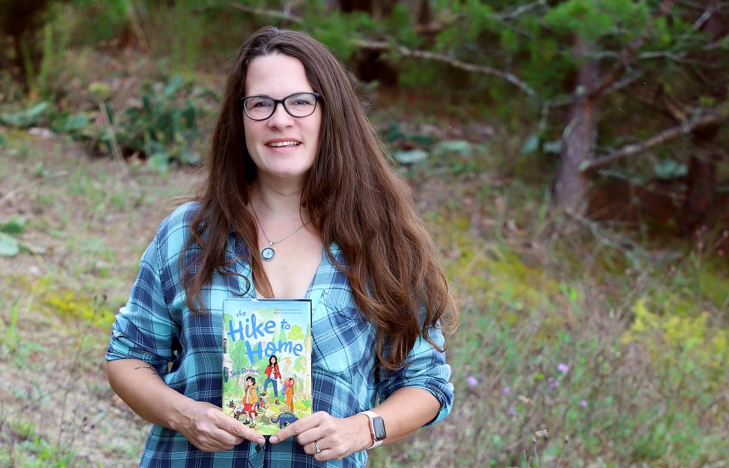 A woman stands and looks at the camera. She is holding a book and the cover of the book says The Hike to Home.