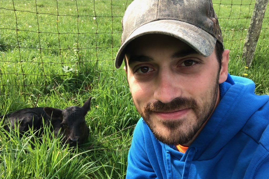 Shayne Turner sits in a green grassy field next to a calf laying near a fence.
