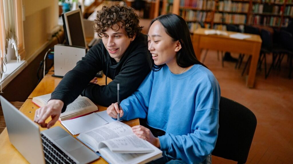Photo of a male and female student writing in a notebook and using a laptop.